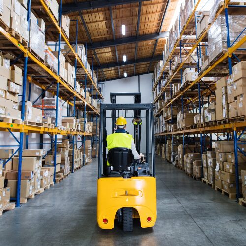 Backside of warehouse employee on a yellow forklift driving through warehouse aisle